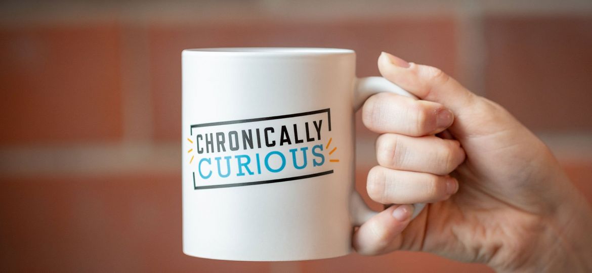 Hand holding white Chronically Curious mug