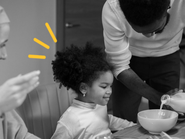 A mother, father and child sit at a table eating breakfast. The father is pouring milk into a cereal bowl for their daughter.